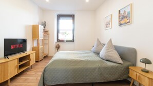 Una camera da letto con un letto, una TV, una cassettiera in legno, una finestra con persiane e un muro decorato da due quadri.