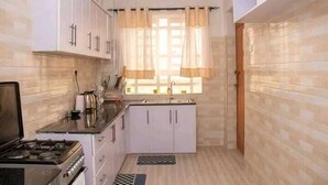 A compact kitchen with white cabinets, a stove, and a window with curtains. - Lux Suites Naxity Apartments Nakuru (Nakuru)