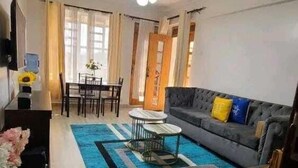 A living room with a sofa, coffee table, and a dining area with chairs and a table. - Lux Suites Naxity Apartments Nakuru (Nakuru)