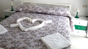 A neatly made bed with a floral bedspread, a heart-shaped towel, and a neatly folded towel on the bed. There is a painting of a coastal landscape on the wall above the bed.