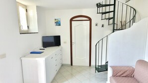 A compact living space with a white wardrobe, a flat-screen TV on a dresser, a door with a wooden frame, and a spiral staircase.