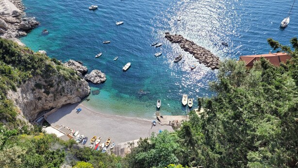 A coastal view with boats, a rocky shoreline, and a beach with lounge chairs.