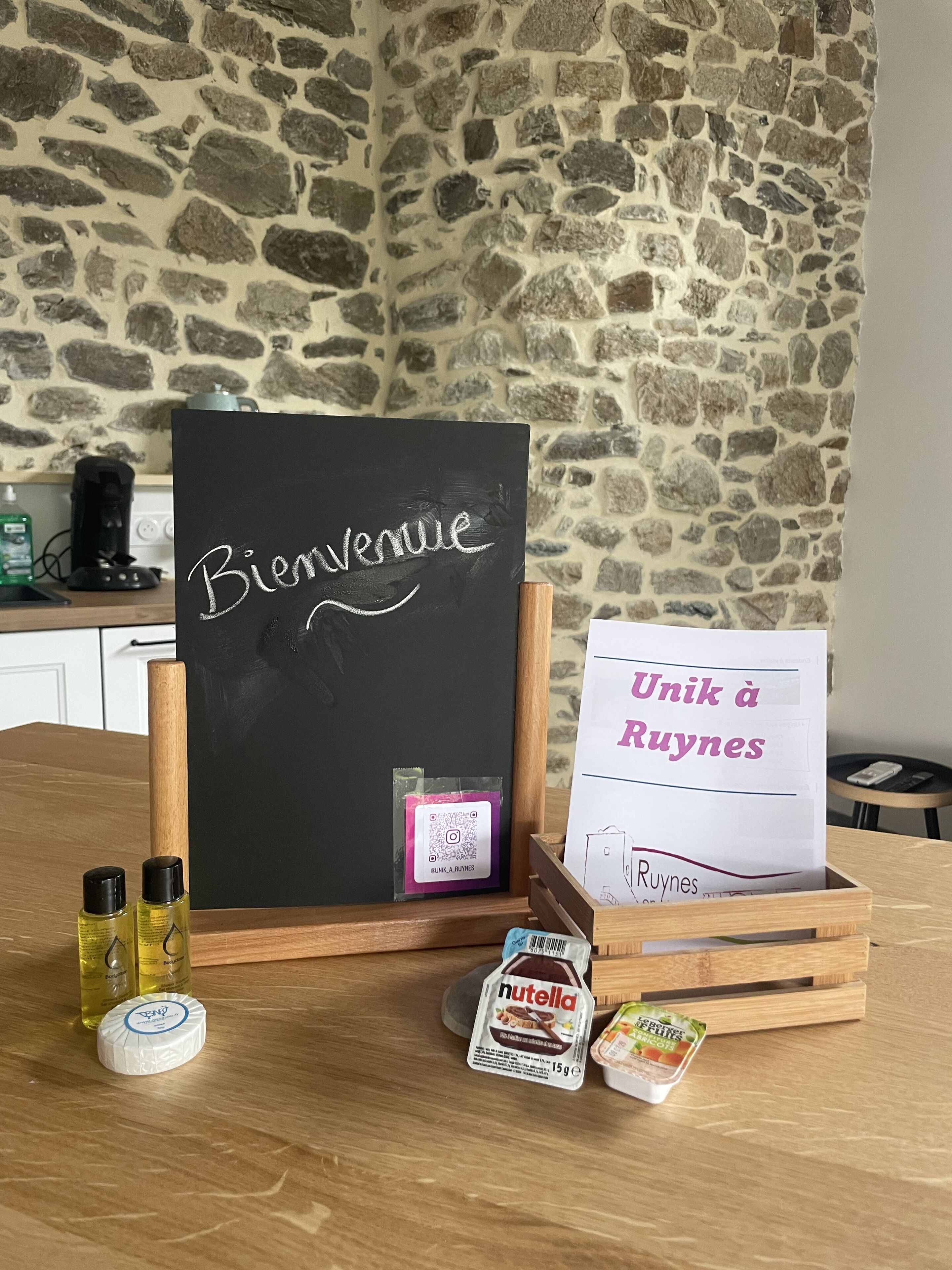 Une table en bois sur laquelle est posé un tableau noir avec l’inscription « Bienvenue », un code QR et un panneau indiquant « Unik à Ruynes ». On y trouve également des bouteilles d’huile, un pot de Nutella et un récipient de yaourt.