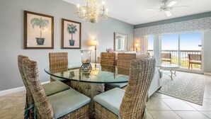 A dining area with wicker chairs, a glass table, and a chandelier. There are framed pictures on the wall and a balcony with a view.