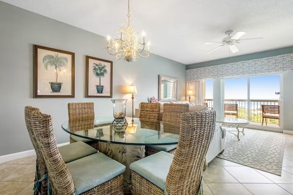 A dining area with wicker chairs, a glass table, and a chandelier. There are framed pictures on the wall and a balcony with a view.