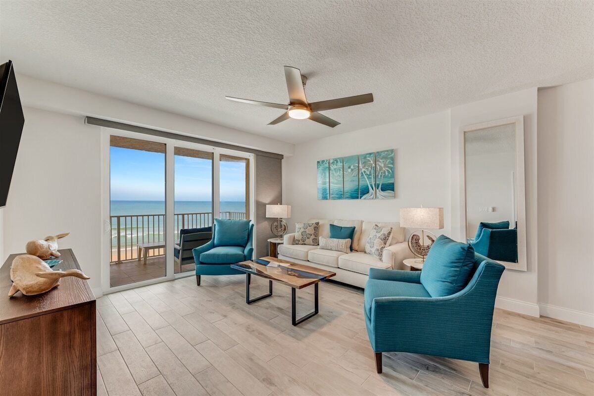 A spacious living room with a large window overlooking the ocean, a ceiling fan, and a modern sofa set.