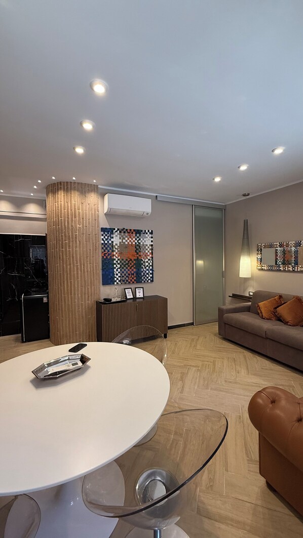 A modern living room with a round glass table, a sofa, and a coffee table. There is a wooden accent wall and a large window.