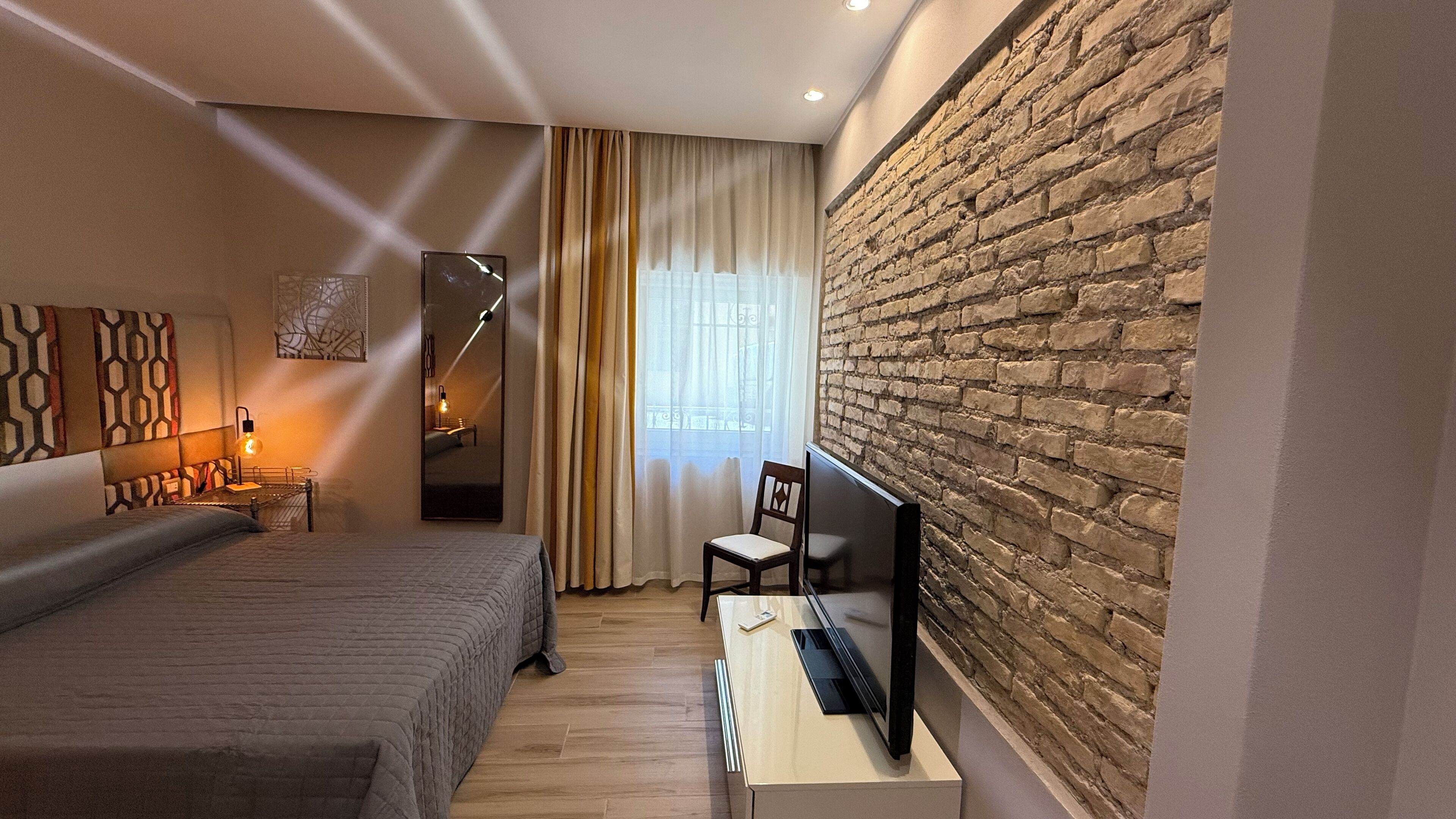 A hotel room with a large bed, a desk with a chair, a television, a mirror, and a brick wall.