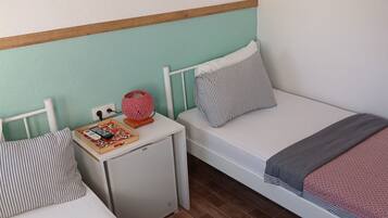 Un dormitorio pequeño y ordenado con dos camas individuales, una mesita de noche blanca y un calentador blanco compacto.