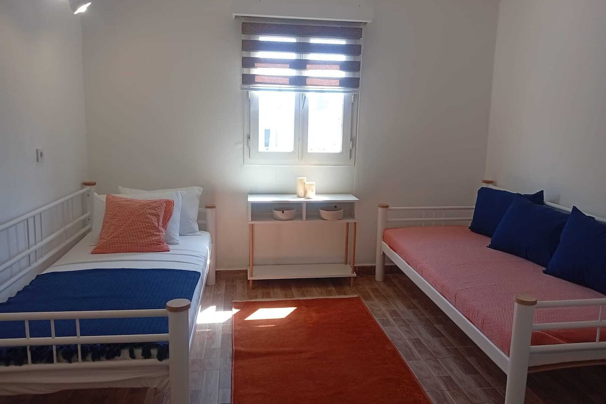 Ein Schlafzimmer mit zwei Betten, einem Fenster mit Jalousien, einem Nachttisch mit Lampe und einem roten Teppich.