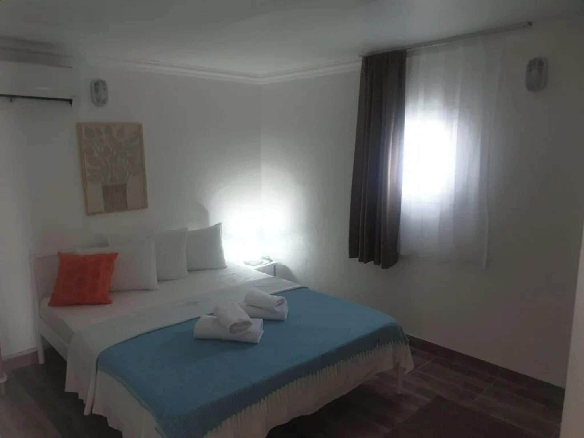 Camera d'albergo con un letto, cuscini, una coperta blu, una finestra con tende e un condizionatore d'aria a parete.