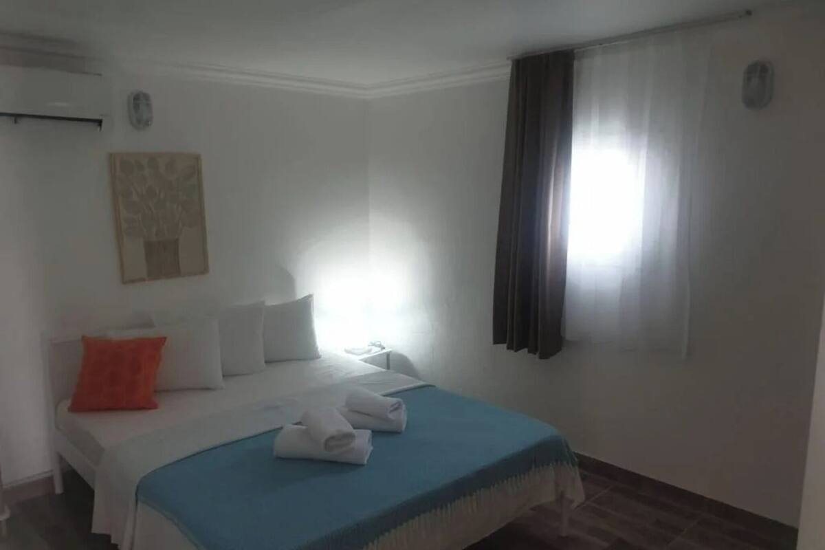 Ein Hotelzimmer mit Bett, Kissen, einer blauen Decke, einem Fenster mit Vorhängen und einer Wand-Klimaanlage.