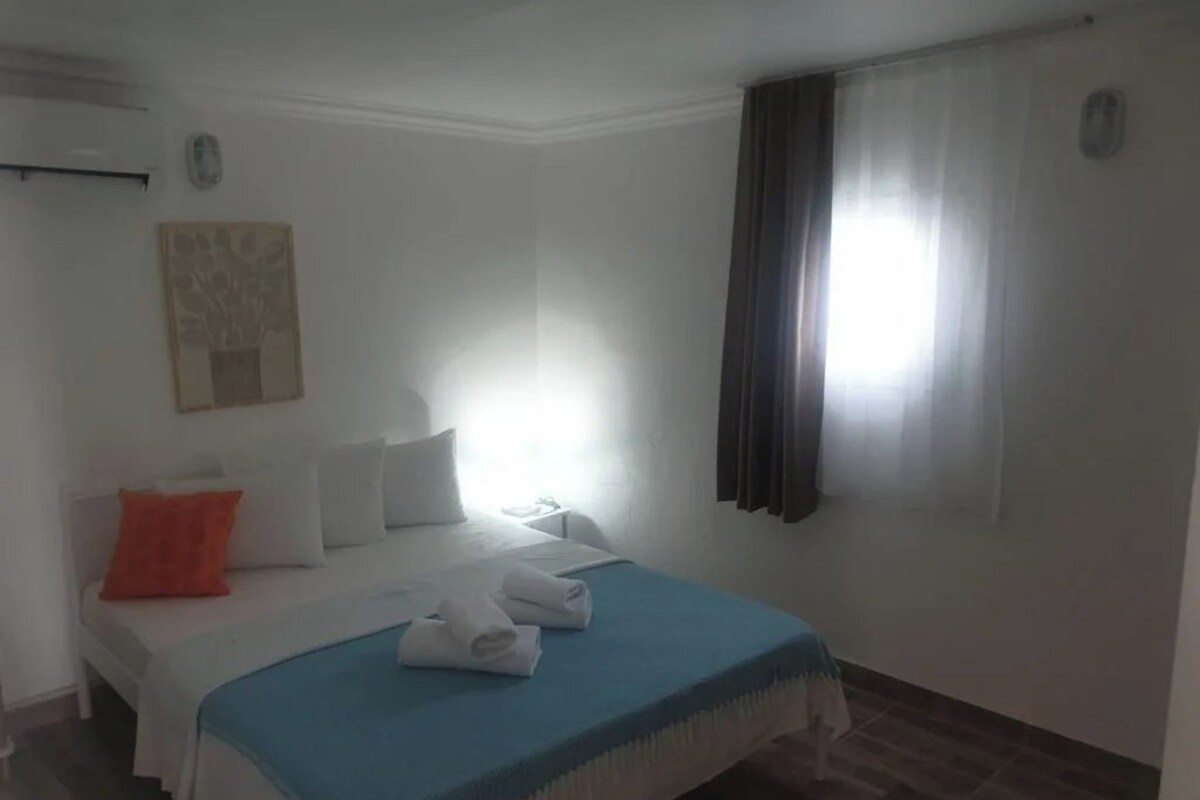 A hotel room with a bed, pillows, a blue blanket, a window with curtains, and a wall-mounted air conditioner.