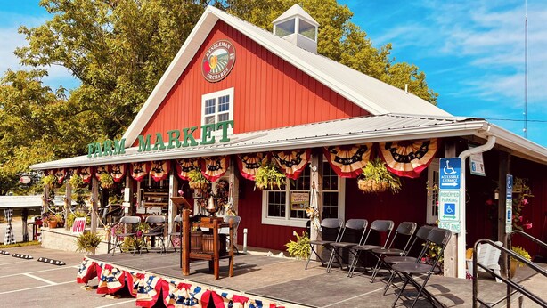 Un bâtiment rouge de style grange, avec une enseigne indiquant « Marché de produits fermiers » et une scène équipée de chaises et d’une table.