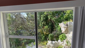 A window with a view of a garden featuring a rock formation, a small pond, and a seating area with chairs.