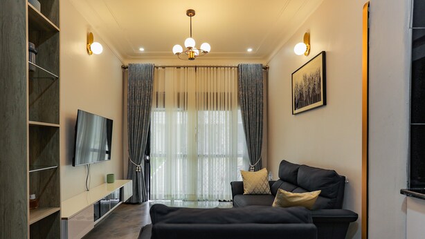 Een moderne woonkamer met een flatscreen televisie, een bank met kussens, een glazen salontafel en een groot raam met gordijnen.