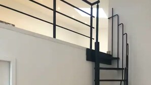 Una escalera moderna con barandilla de metal negro, paredes blancas y una puerta a la izquierda.