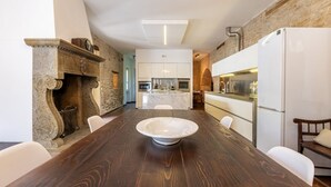 A dining area with a large wooden table, white chairs, a stone fireplace, and a kitchen in the background. - Villa Roncaglia by MMega (Fratte Rosa)