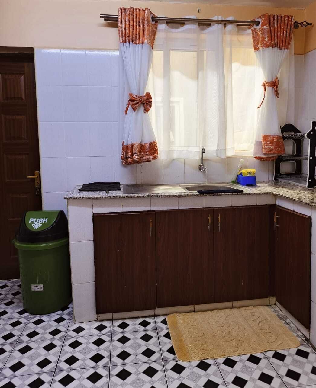 A kitchen with a tiled floor, a sink, cabinets, and a trash bin.