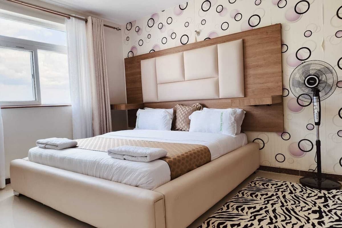 Una habitación de hotel moderna con una cama grande, un cabecero, un ventilador de pie y una alfombra estampada.