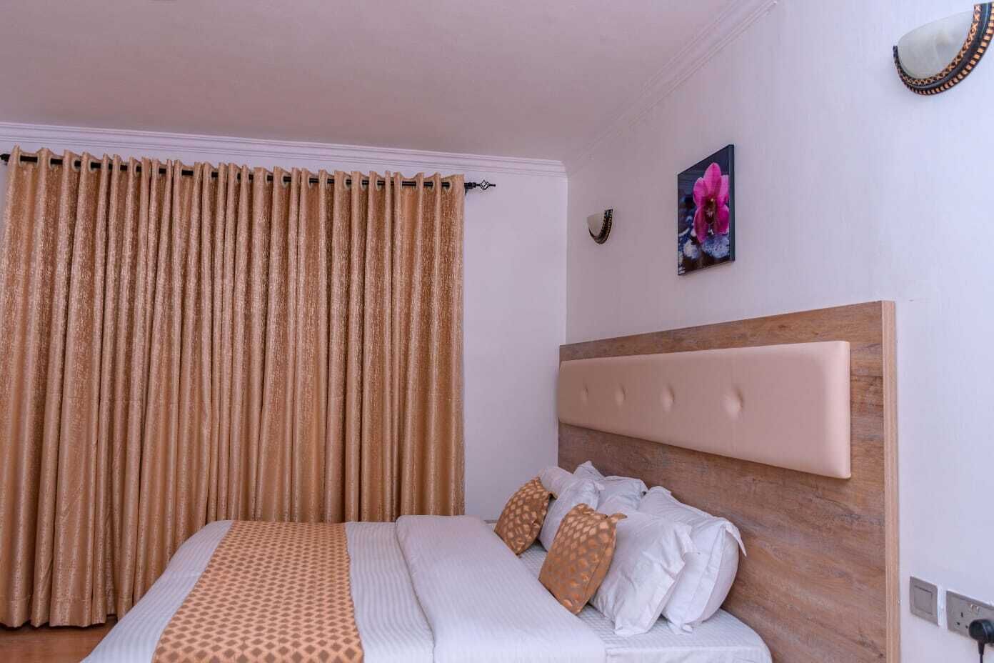 A hotel room with a large bed, beige curtains, a wooden headboard, and a small decorative item on the wall.