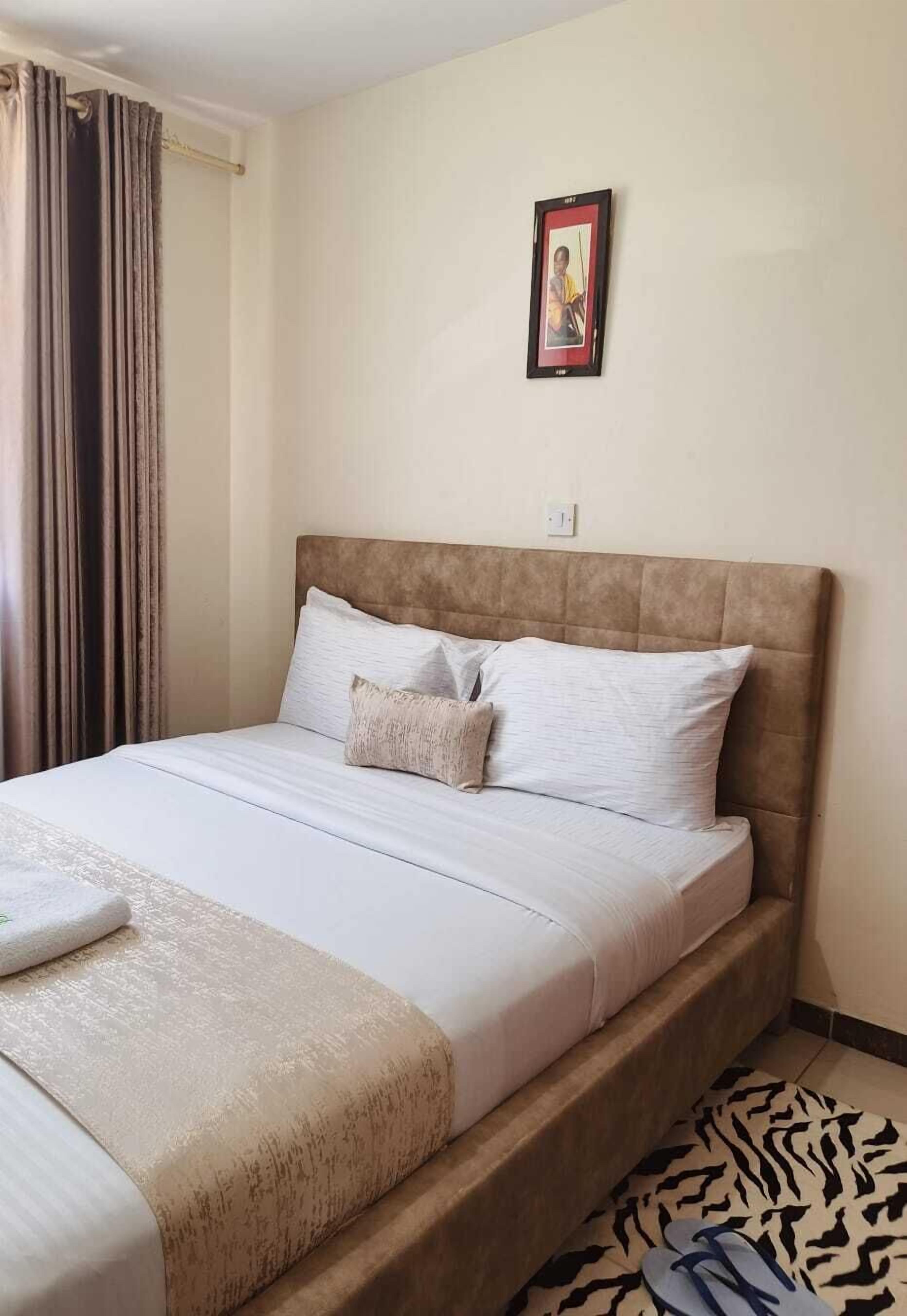 A neatly made bed with a headboard, a framed picture on the wall, and a pair of slippers on the floor.