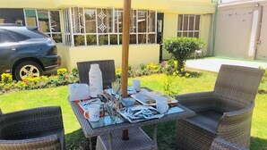 Eine Terrasse mit Korbmöbeln und einem Essplatz, umgeben von einem Garten und einem Gebäude mit Fenstern.