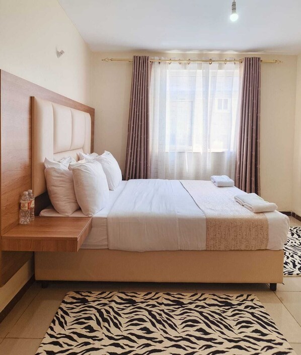A neatly made bed with white linens, a wooden headboard, and a zebra-patterned rug on the floor. - Lux Suites Havan Furnished Apartments (Nakuru)