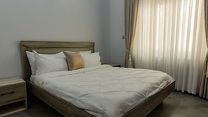 Una cama bien hecha con cabecera de madera, una mesita de noche con cajón y una ventana con cortinas translúcidas.