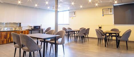 A modern conference room with multiple tables and chairs, a kitchenette, and a projection screen on the wall.