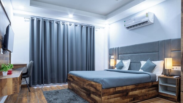 A modern bedroom with a wooden bed frame, a desk with a chair, a TV mounted on the wall, and a large window with curtains.
