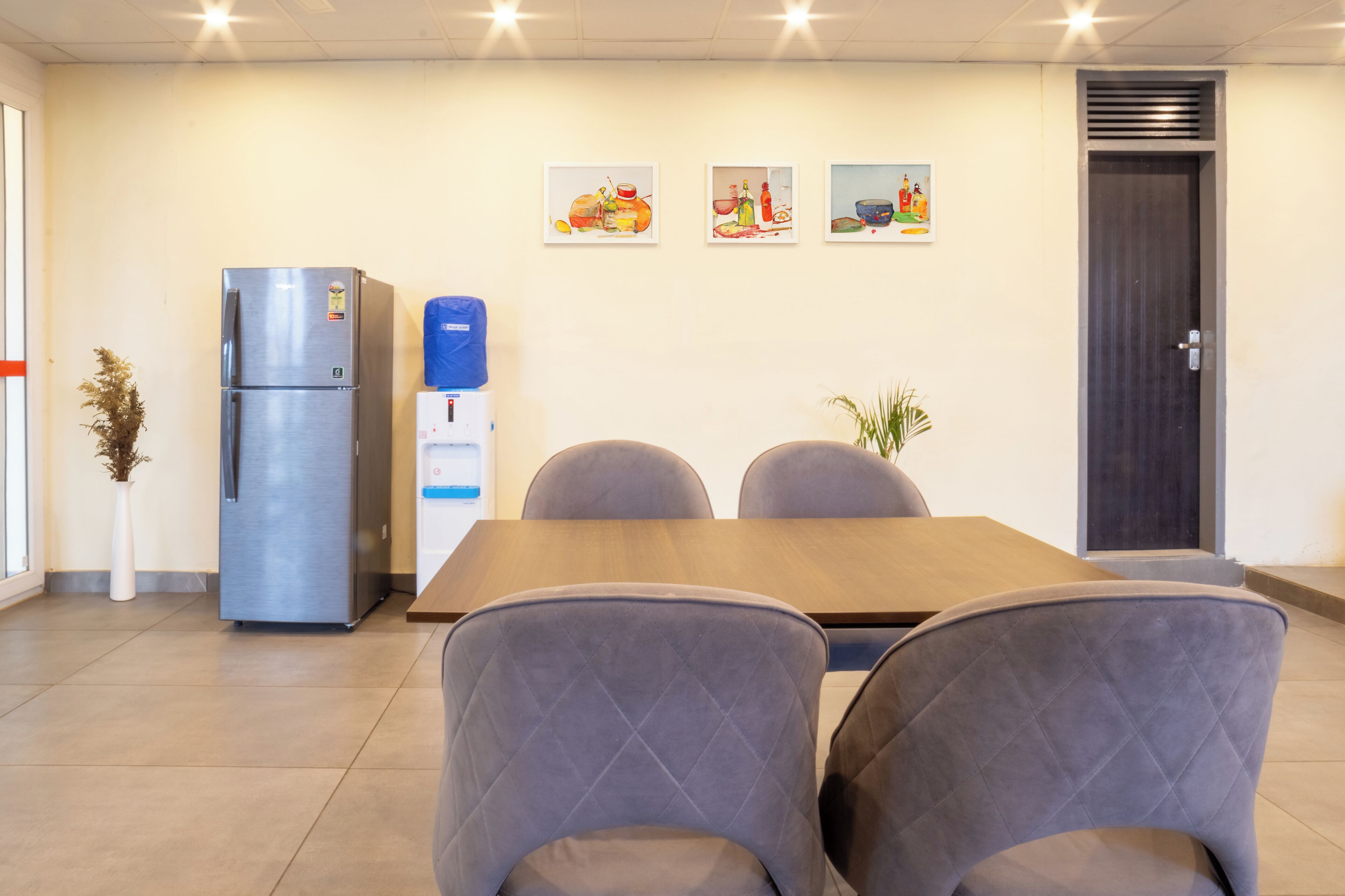 A modern meeting room with a wooden table, four grey upholstered chairs, a stainless steel refrigerator, and three framed artworks on the wall.