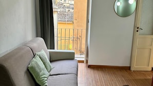 A room with a sofa, a wooden floor, a door, and a view of buildings through an open door.