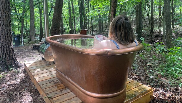 A person relaxing in a large, brown bathtub outdoors surrounded by trees.