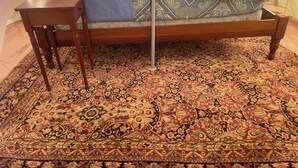 A bed with a patterned bedspread, a blue blanket, and a grey throw. There is a wooden nightstand with a lamp. The room has a large, ornate carpet.