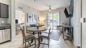 Een moderne keuken met een inbouwvaatwasser, een eethoek met een ronde tafel en stoelen, een woonkamer met een bank en een televisie, en een groot raam met uitzicht op buiten.