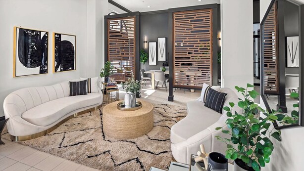 Een moderne woonkamer met een gebogen witte bank, een ronde salontafel en een grote spiegel. Twee houten roomscheidingen met planten, en er is een eethoek zichtbaar op de achtergrond.