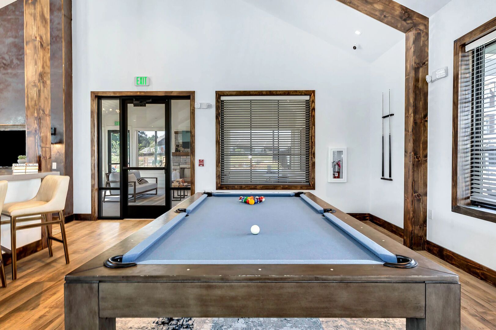 A pool table in a room with wooden beams, a door with an exit sign, and a window with blinds.