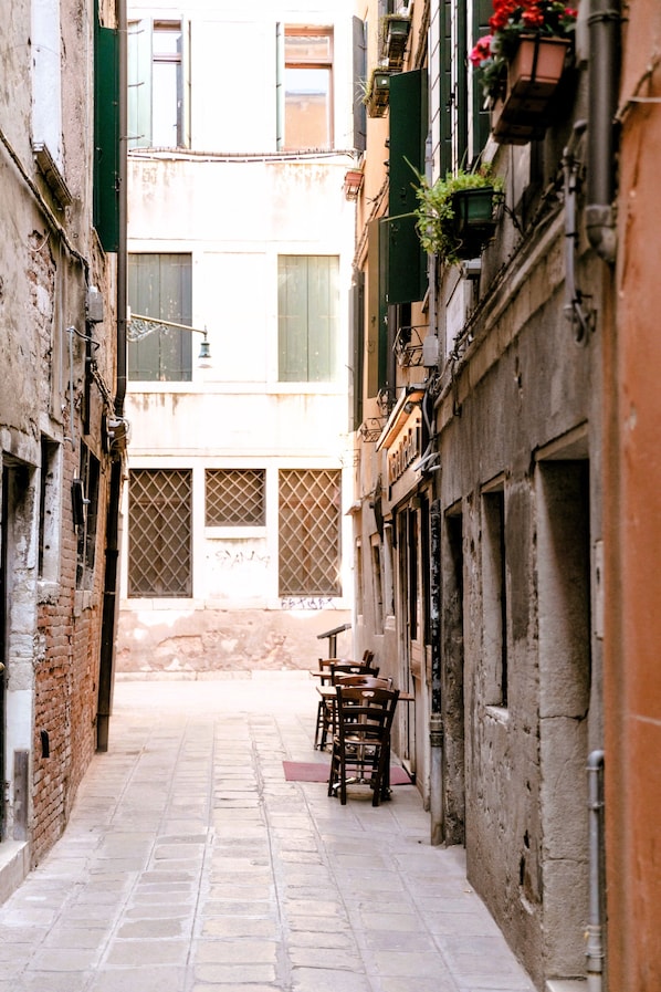 Un estrecho callejón con pavimento de piedra, mobiliario de madera y edificios con contraventanas verdes y macetas con flores.
