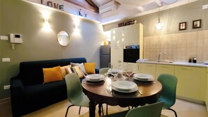 A modern living room with a dark blue sofa, green chairs, and a dining table set for four. A kitchen with a sink and stove is visible in the background. - La Serenissima: 2BR Castello Apartment, Private Roof Terrace, 3 min to San Marco (Venezia)