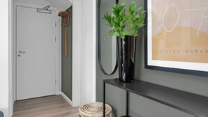 A hallway with a door, a mirror, a potted plant, a framed artwork, and a woven stool.