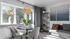A modern living room with a round table, two chairs, a sofa, a large window with curtains, a hanging light fixture, and a bookshelf.