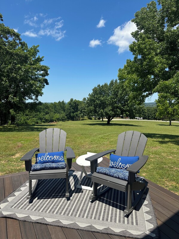 Zwei Adirondack-Stühle mit 'welcome'-Kissen auf einer Holzterrasse mit einem gewebten Teppich.
