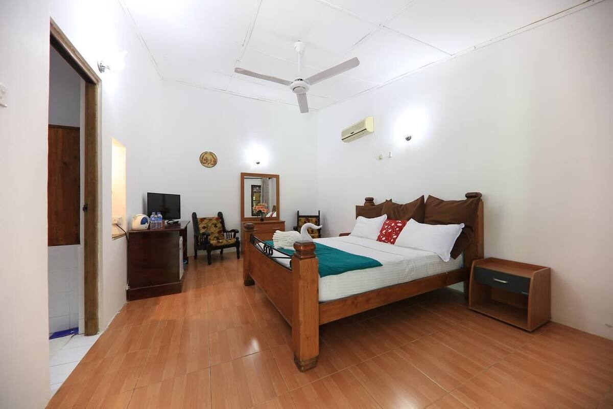 Een slaapkamer met een houten bed, een nachtkastje, een bureau met stoel, een plafondventilator en een wandairconditioner.