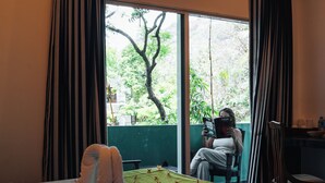Ein Zimmer mit einem Bett, einem grünen Tisch mit roten Kreuzen, einem Stuhl und einer Person, die am Fenster ein Buch liest und dabei einen Blick auf Bäume hat.