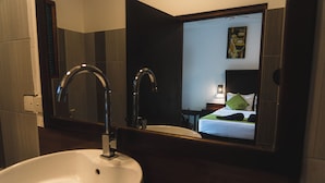 Een badkamer met een witte wastafel, twee kraanwerk en een spiegel die een bed en een schilderij weerspiegelt.