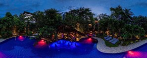 Vista nocturna de un complejo turístico con un amplio área de piscina, sillas de descanso y una terraza de madera.