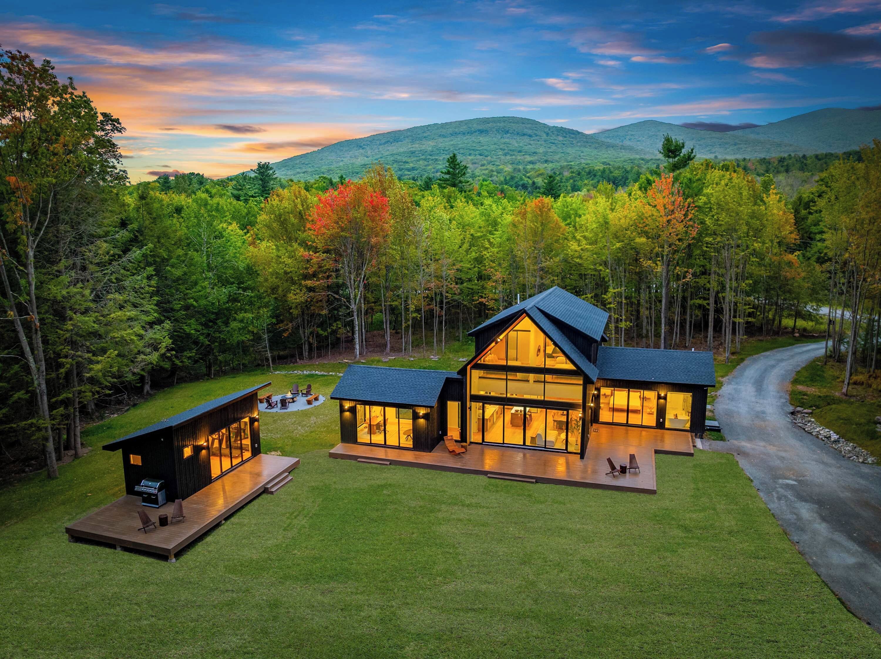 A modern cabin with a large deck, surrounded by a forest and a mountain backdrop.