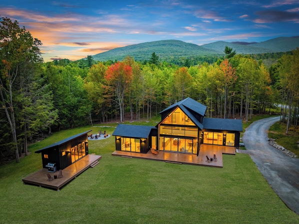 A modern cabin with a large deck, surrounded by a forest and a mountain backdrop.