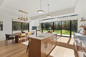 A modern kitchen with a large island, dining area, and large windows offering a view of a forest.